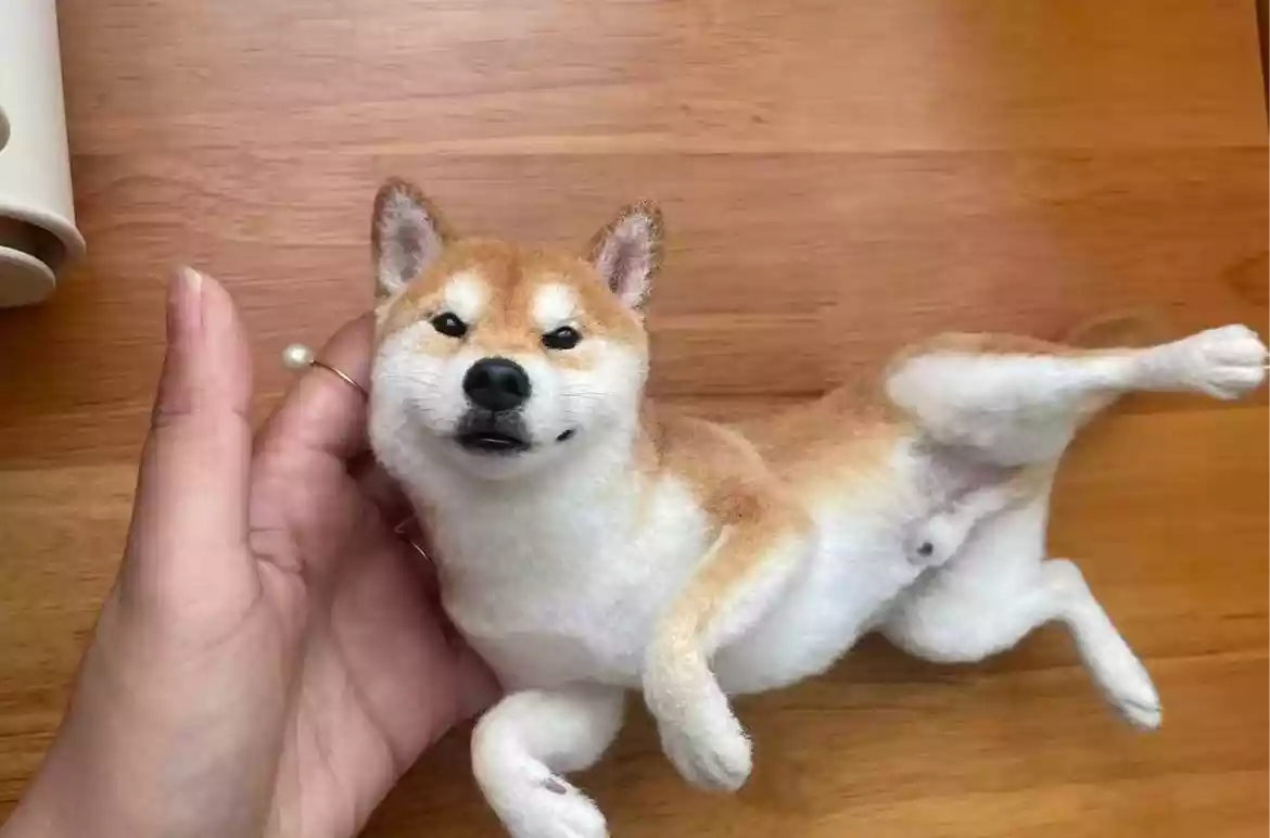 Hand holding a custom 3D needle-felted Shiba Inu replica on a wooden floor.